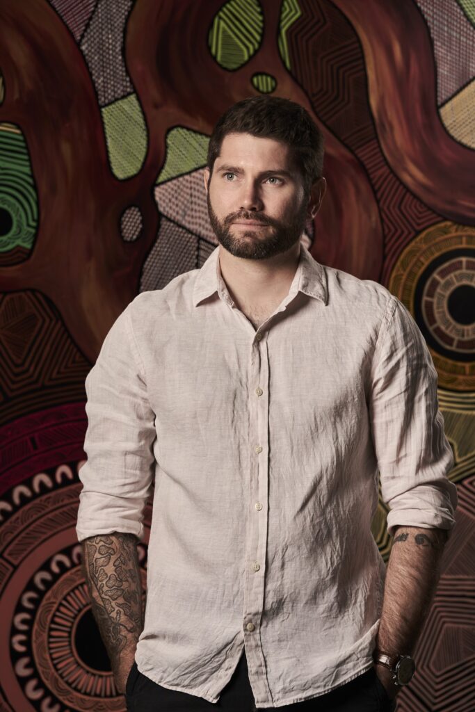 Troy Firebrace standing in front of his large aboriginal artwork
